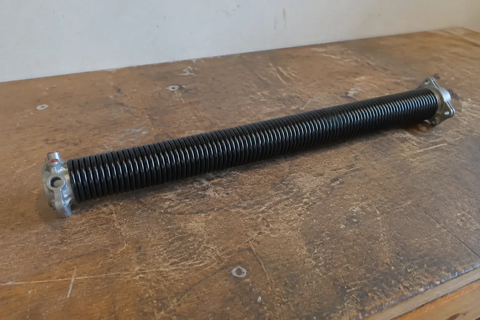 Detached black coil spring with metal end fittings on a workbench. — Torsion Spring Replacement, Mississauga